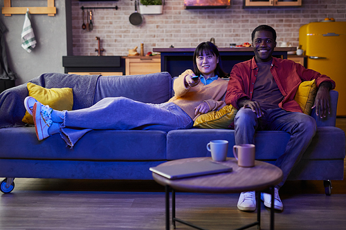 Full length portrait of young couple watching TV while relaxing on couch in cozy home interior, copy space