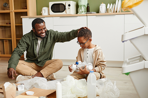 Full length portrait of happy African-American father and son sorting plastic and paper at home for waste recycling, copy space