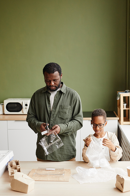 Vertical portrait of African-American Father and son preparing household waste for recycling and cutting out labels