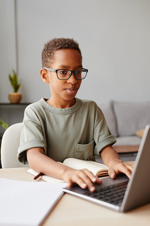 Vertical portrait of smiling African-American boy wearing glasses using laptop while studying at home, homeschooling concept