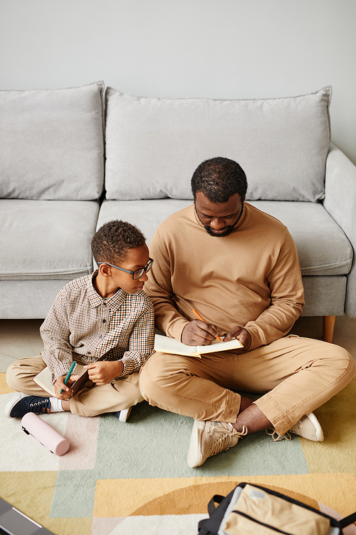 Vertical full length portrait of African-American father and son sitting on floor while doing homework together
