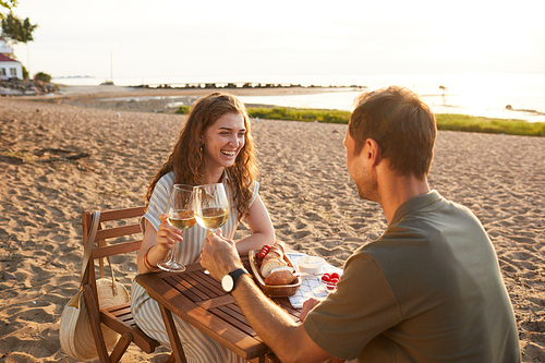 Portrait of happy young couple drinking wine at picnic outdoors while enjoying romantic date on beach, copy space