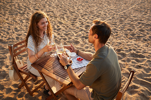 High angle portrait of happy young couple drinking wine at picnic outdoors while enjoying romantic date on beach, copy space