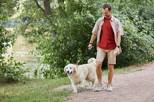 Full length portrait of smiling man walking dog outdoors in Summer at green park, copy space