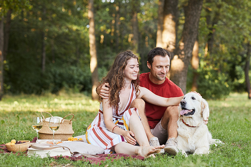 Full length portrait of young couple with dog enjoying picnic outdoors on green grass, copy space