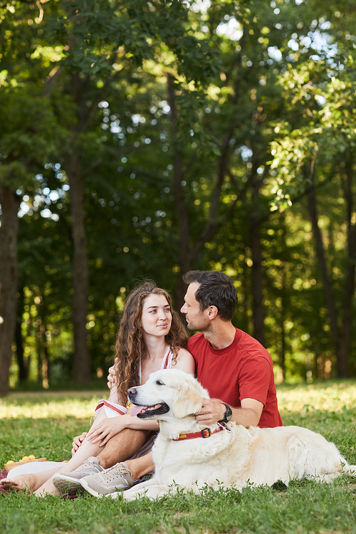 Vertical full length portrait of young couple with dog enjoying picnic outdoors on green grass