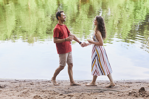 Full length portrait of happy couple holding wine glasses while enjoying romantic date by water outdoors, copy space