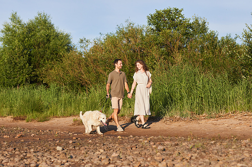 Wide angle portrait of happy couple with dog walking together by water in sunlight, copy space