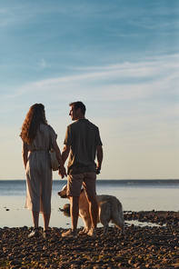 Back lit full length portrait of happy couple with dog together by water at sunset