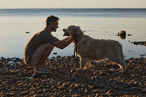 Back lit full length portrait of happy man playing with dog by water at sunset, copy space