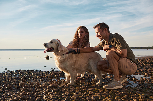 Back lit full length portrait of happy young couple playing with dog by water at sunset, copy space