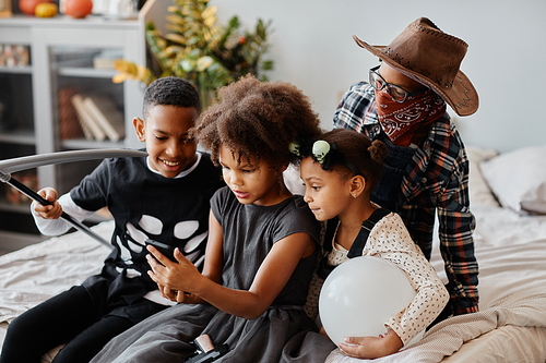 Group of African-American kids wearing Halloween costumes at home and using smartphone