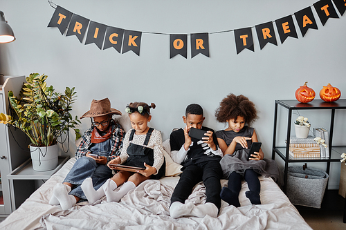 High angle portrait of four African-American children using gadgets in bed under Halloween decorations at home