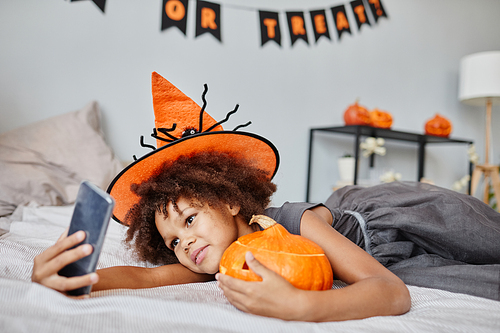 Portrait of cute girl wearing Halloween costume and taking selfie while lying on bed at home, copy space