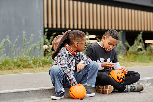 Full length portrait of two African-American boys holding Halloween buckets while sitting on curb outdoors during trick or treating, copy space