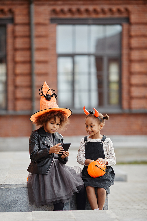 Vertical portrait of two African-American girls wearing Halloween costumes outdoors and using smartphone, copy space