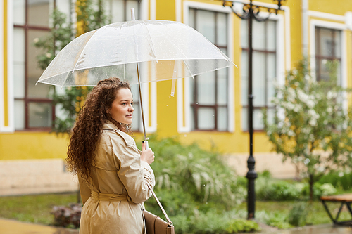 Horizontal shot of stylish young Caucasian woman with long curly hair standing outdoors under transparent umbrella on rainy day looking away