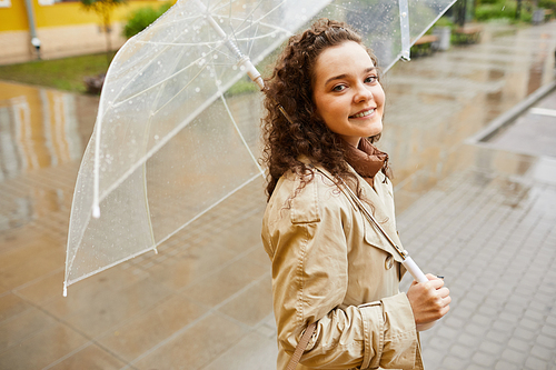 Horizontal high angle medium portrait of beautiful young Caucasian woman wearing beige trench coat standing outdoors under umbrella on rainy day smiling at camera