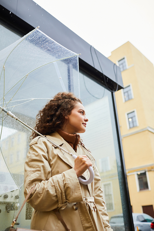 Vertical low angle shot of young woman wearing beige trench coat standing under umbrella on bus stop looking away