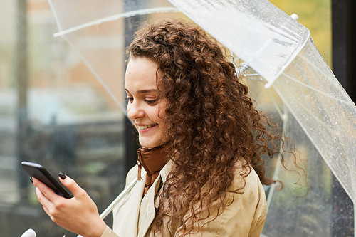 Horizontal medium close-up portrait of Caucasian woman with curly hair standing outdoors under umbrella on rainy day using Internet on smartphone