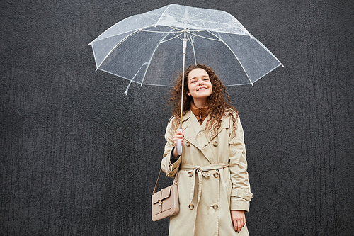 Horizontal shot of attractive young woman wearing trench coat holding umbrella standing outdoors against gray wall looking at camera