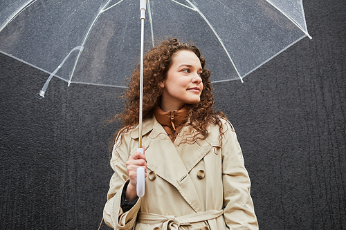 Horizontal medium shot of young woman wearing trench coat holding transparent umbrella standing outdoors against gray wall looking away