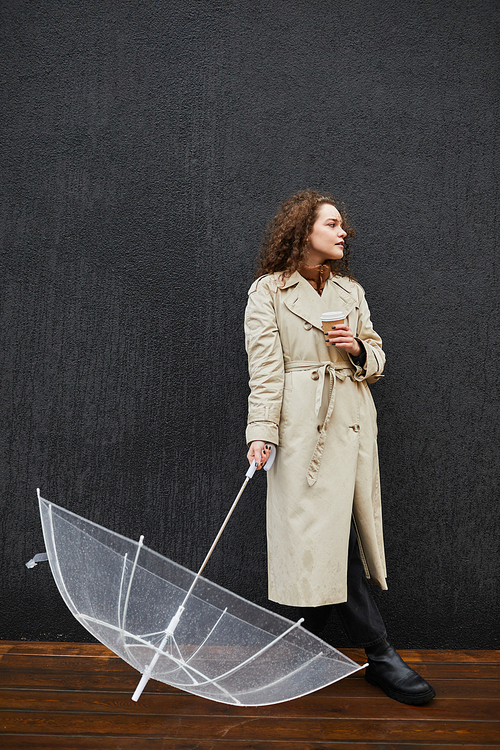 Vertical full shot of young woman wearing trench coat holding transparent umbrella and cup of coffee standing outdoors against gray wall looking away