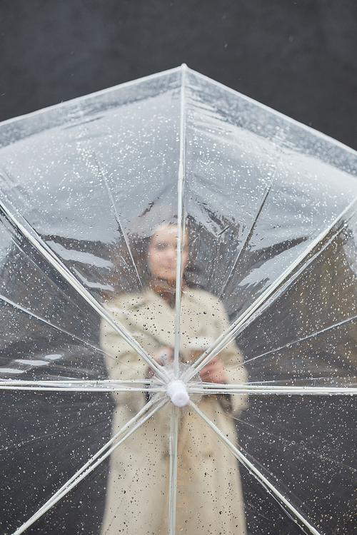 Vertical through transparent umbrella shot of young woman wearing trench coat standing outdoors against gray wall looking at camera