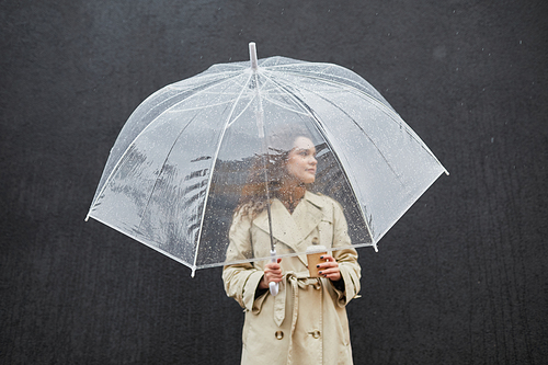 Horizontal medium shot of modern young woman wearing trench coat holding transparent umbrella and cup of coffee standing outdoors against dark gray wall looking away