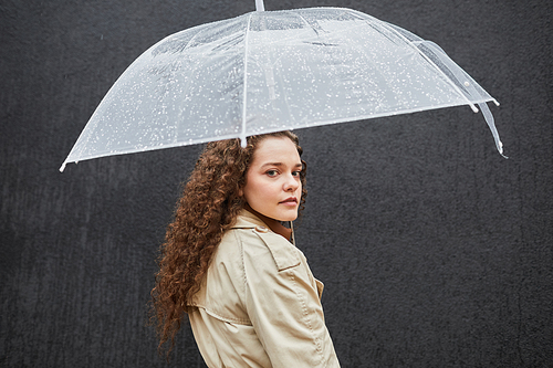 Horizontal medium shot of beautiful young woman with curly hair wearing trench coat standing under umbrella outdoors against dark gray wall looking at camera
