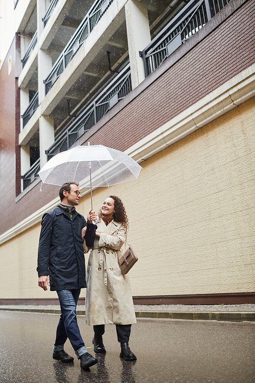 Vertical long shot of stylish Caucasian man and woman spending time together outdoors walking under umbrella