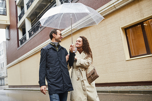 Horizontal medium shot of modern Caucasian man and woman in love enjoying time together outdoors walking under umbrella and smiling