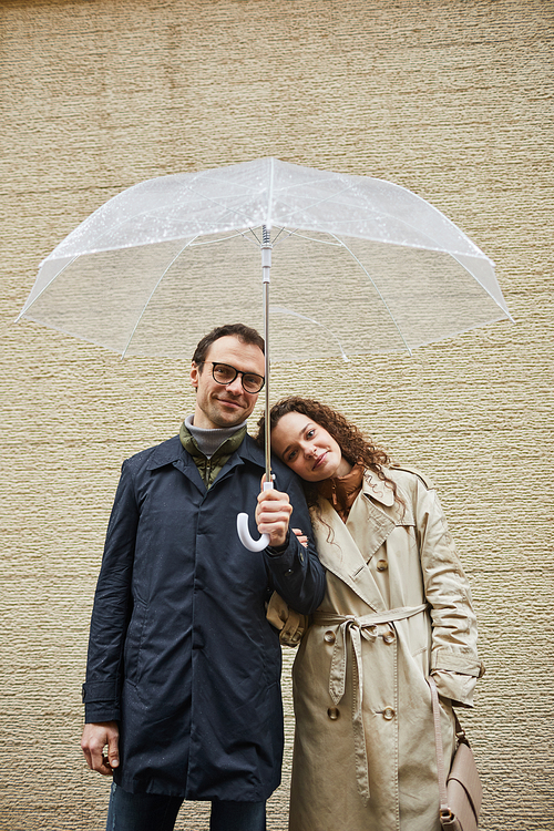 Vertical shot of happy couple spending time together outdoors standing against beige building wall under umbrella looking at camera