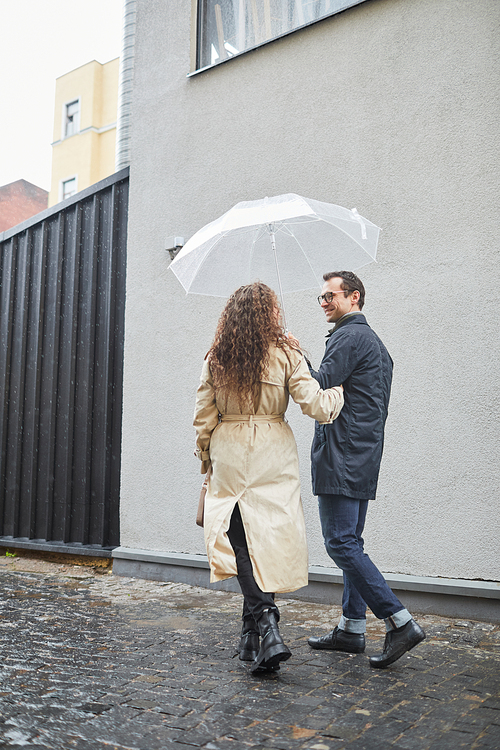 Vertical medium shot of Caucasian man spending time with his girlfriend outdoors walking on rainy day under umbrella