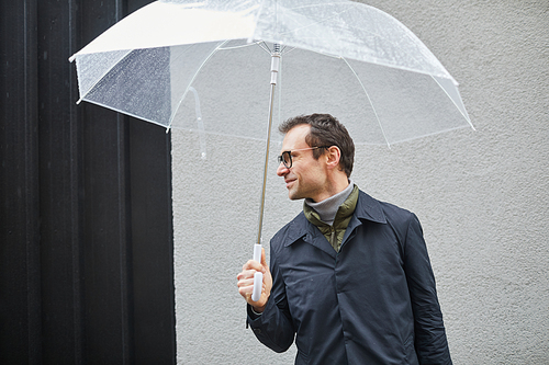 Horizontal medium shot of handsome Caucasian man standing outdoors under transparent umbrella looking away