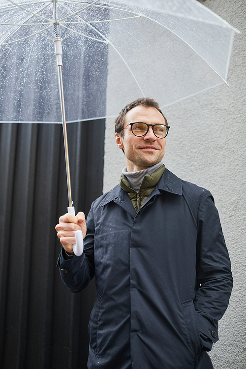 Vertical medium shot of stylish Caucasian man wearing eyeglasses standing outdoors under transparent umbrella looking away