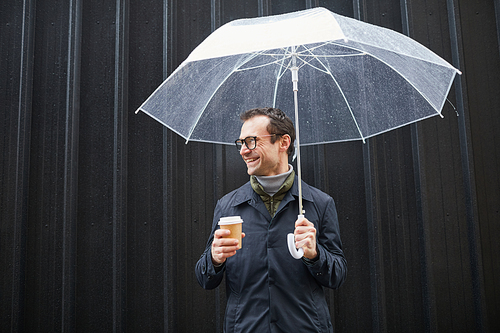 Horizontal medium shot of stylish Caucasian man wearing eyeglasses standing outdoors under transparent umbrella holding coffee cup, looking away and smiling