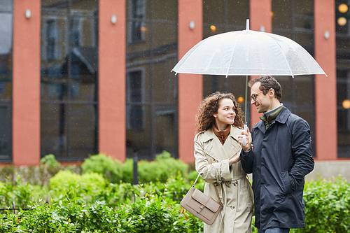 Horizontal medium shot of Caucasian man spending time with his girlfriend outdoors walking on rainy spring day under umbrella