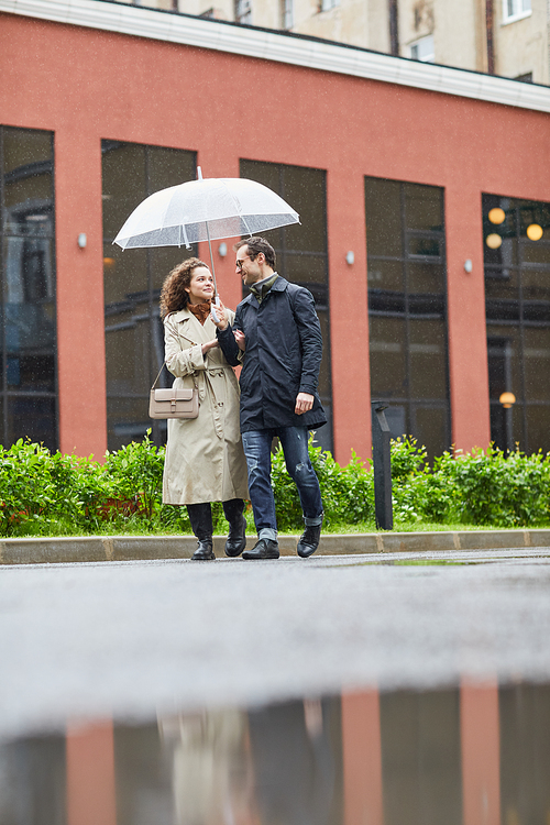 Vertical long shot of Caucasian man and woman walking along street on rainy spring day under umbrella