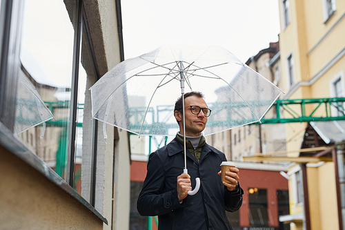 Horizontal medium shot of stylish Caucasian man wearing eyeglasses walking along street under umbrella holding coffee cup, looking away