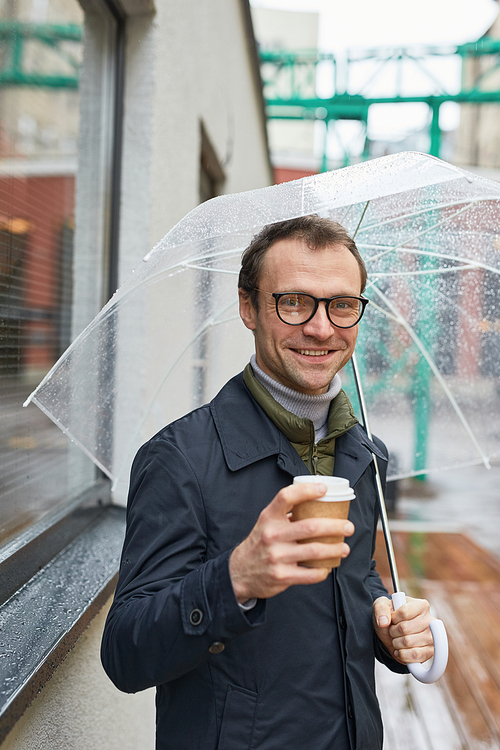 Vertical medium shot of handsome Caucasian man wearing eyeglasses walking along street under umbrella holding coffee cup, looking at camera