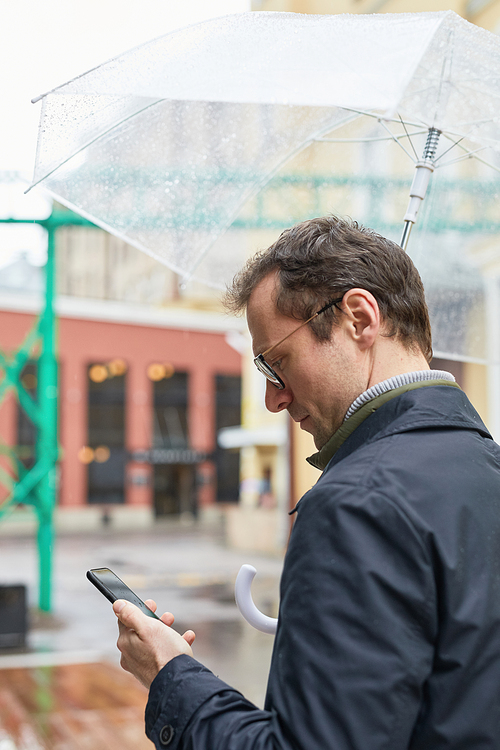 Vertical medium shot of modern Caucasian man wearing eyeglasses spending time outdoors standing under umbrella texting something on smartphone