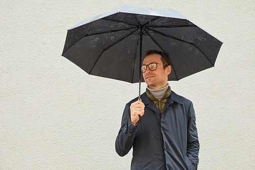 Horizontal medium shot of handsome Caucasian man wearing eyeglasses standing outdoors under umbrella looking away