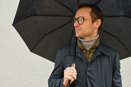 Horizontal chest up shot of handsome Caucasian man wearing eyeglasses and black raincoat standing outdoors under umbrella looking away