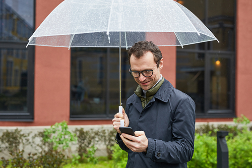 Horizontal medium shot of joyful Caucasian man wearing eyeglasses spending time outdoors standing under umbrella in rain surfing Internet on smartphone