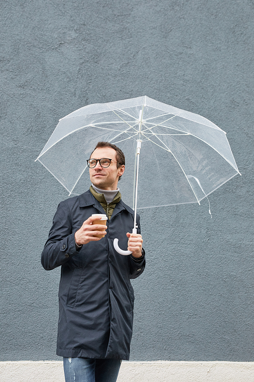 Vertical medium long shot of stylish Caucasian man wearing eyeglasses standing under umbrella with cup of coffee looking away