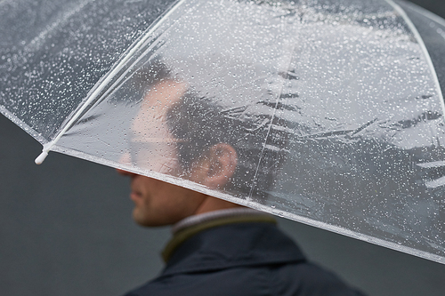 Medium close-up selective focus shot of mature Caucasian man wearing eyeglasses standing under transparent umbrella on rainy day
