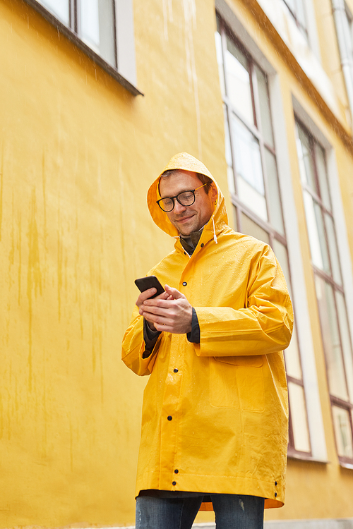 Vertical low angle shot of mature Caucasian man wearing yellow raincoat walking in rain using smartphone