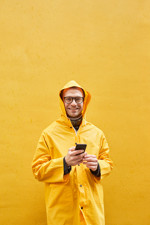 Vertical portrait of modern Caucasian man wearing yellow raincoat standing against yellow wall holding smartphone looking at camera