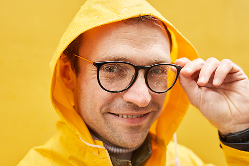 Close-up portrait of stylish Caucasian man wearing yellow raincoat standing against yellow wall taking off eyeglasses looking at camera